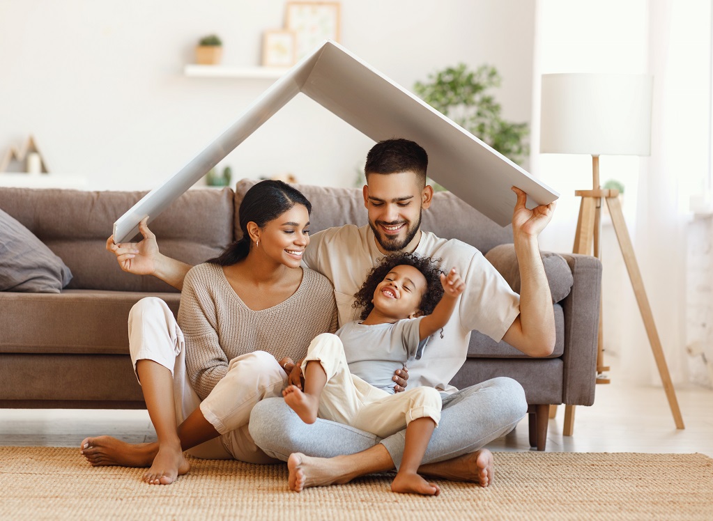 Delighted diverse parents and mixed race son sitting under roof and smiling near sofa in cozy living room at hom